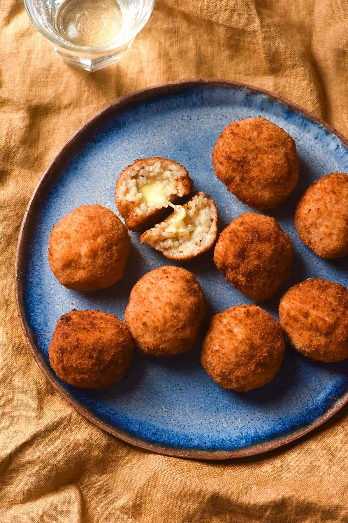 An aerial image of gluten free arancini on a blue ceramic plate atop a mustard linen tablecloth. One arancino has been split open, revealing the cheesy innards. A glass of water sits in the top left of the image. 