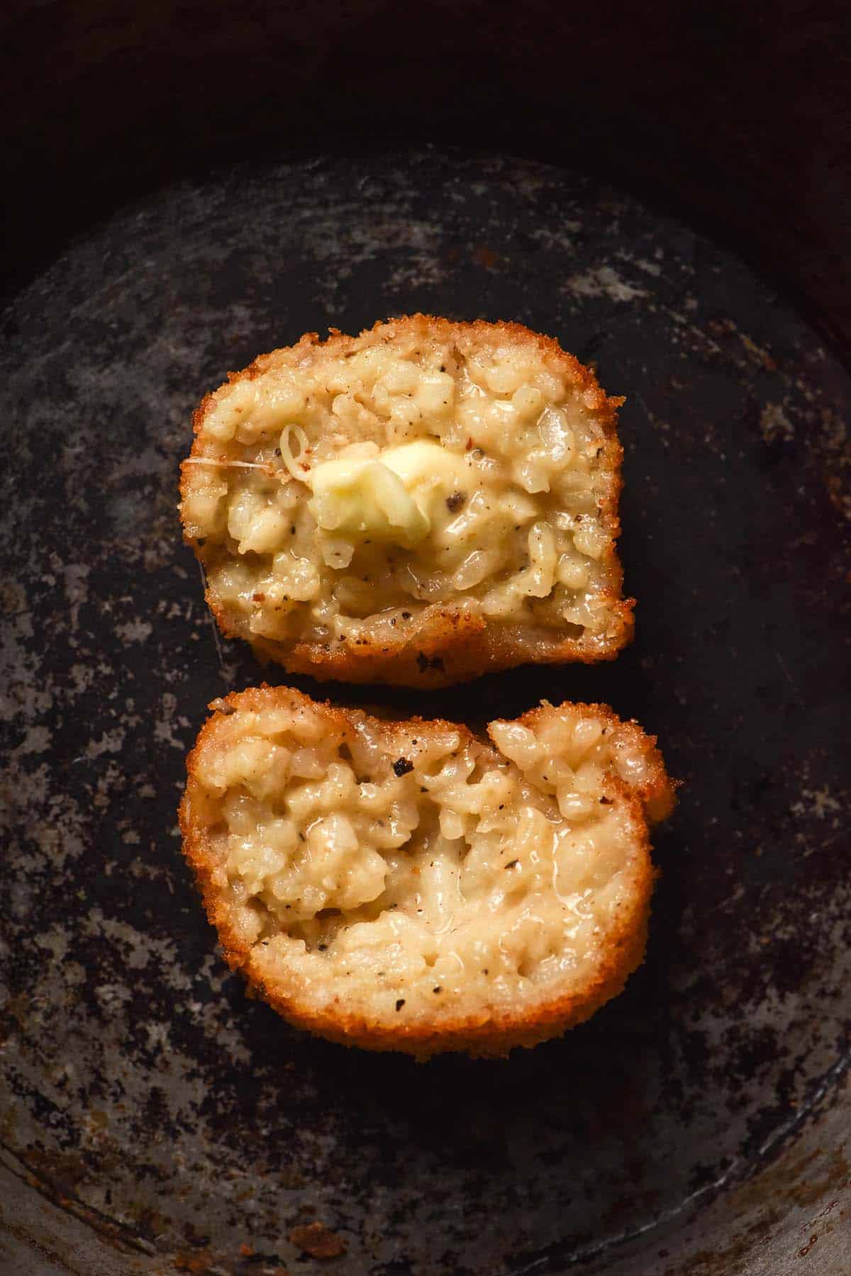 An aerial image of an arancini ball that sits atop a white rustic skillet. The arancini has been torn in half, revealing the cheesy centre inside. 