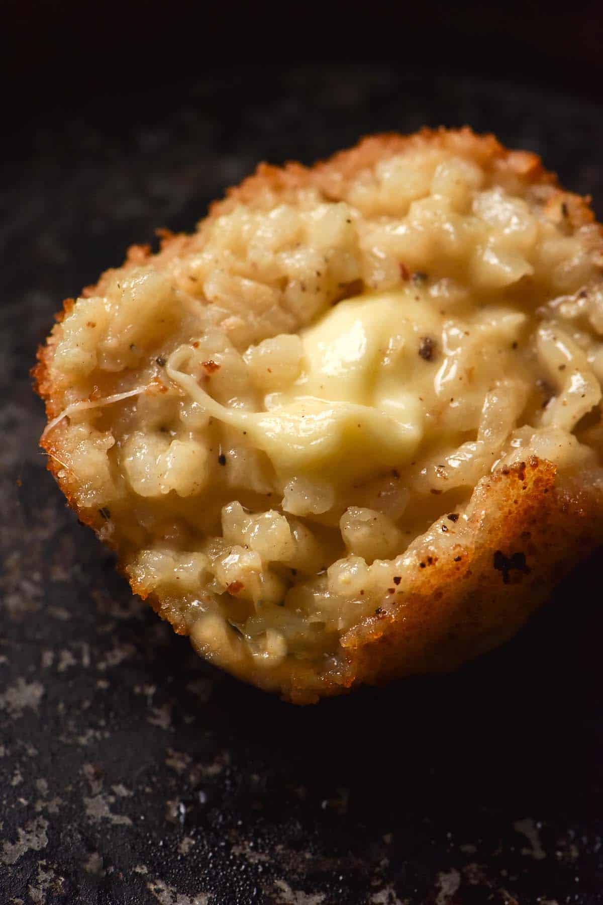 A close up macro image of a gluten free arancino ball that has been torn open to revealing the cheesy innards. It sits on a dark grey oven tray. 