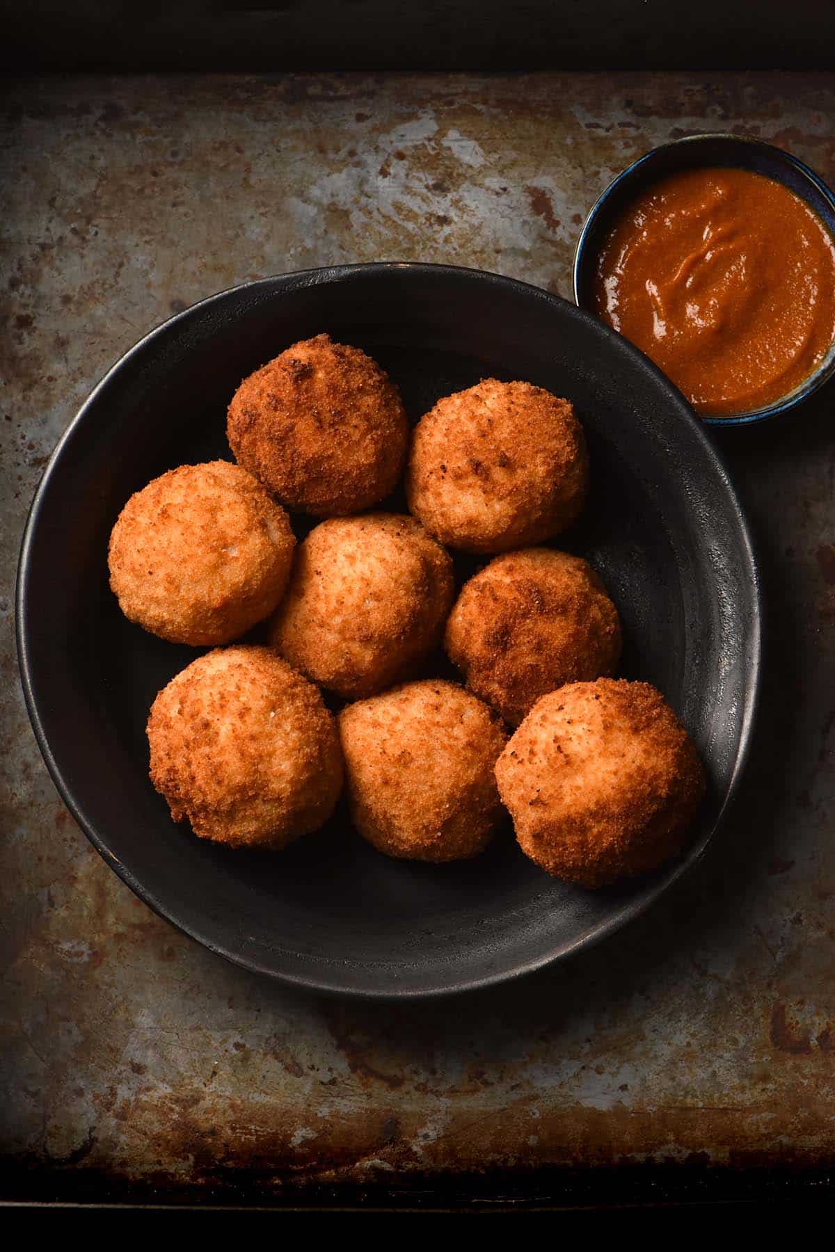 An aerial image of gluten free arancini in a dark grey ceramic bowl atop a mottled grey backdrop. A small bowl of marinara sits to the top right of the arancini. 