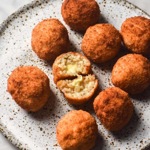 A sunlit view of a white speckled ceramic plate topped with gluten free arancini. One arancini has been torn in half, revealing the cheesy centre. A glass of water sits to the top left of the image.