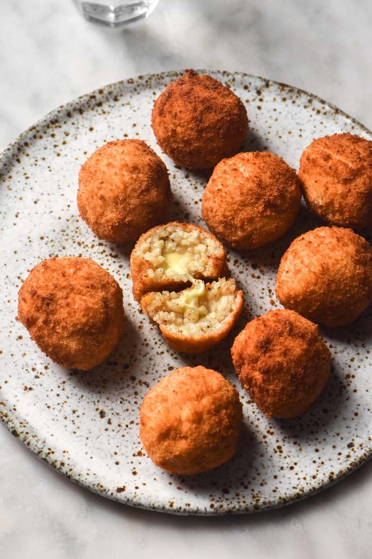 A sunlit view of a white speckled ceramic plate topped with gluten free arancini. One arancini has been torn in half, revealing the cheesy centre. A glass of water sits to the top left of the image.