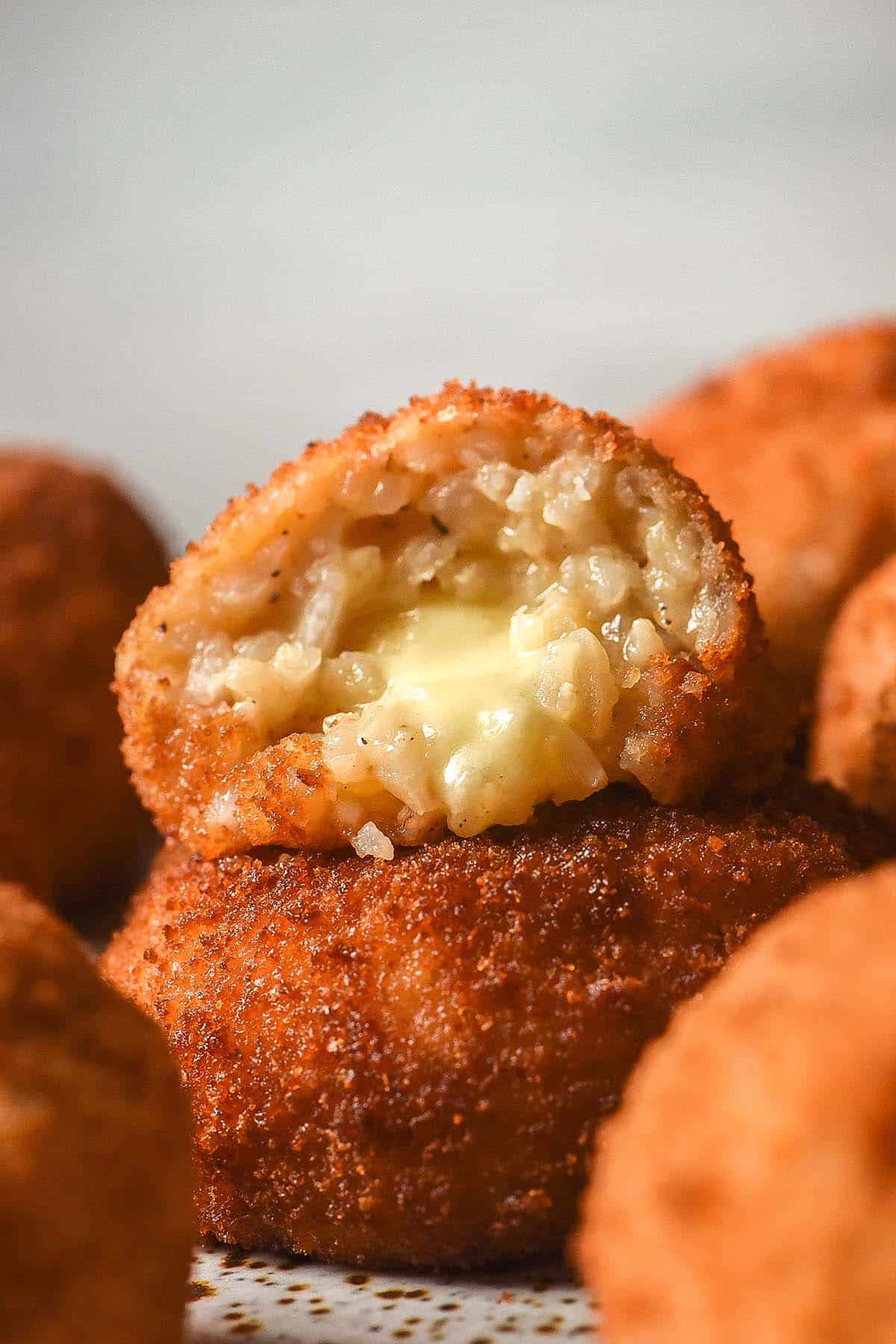 A side on image of gluten free arancini on a white plate against a white background. The arancini are stacked and the top arancino has been pulled apart, revealing the cheesy inner core.