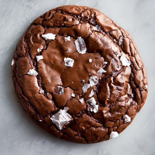 An aerial close up image of a gluten free brownie cookie topped with sea salt flakes on a white marble table.