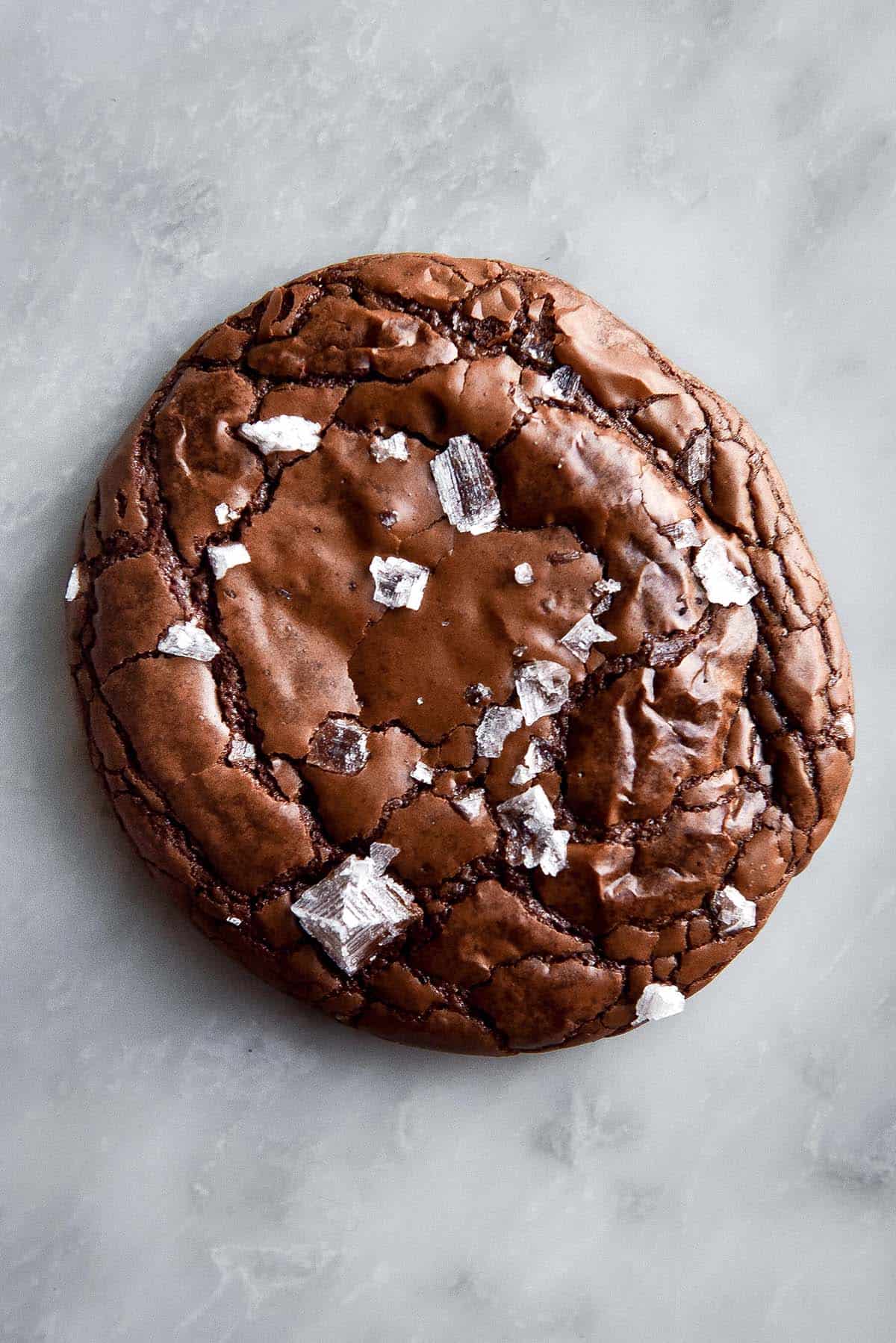 A close up macro image of a gluten free brownie cookie topped with sea salt flakes. The brownie cookie is fudgy and crinkled and sits atop a white marble table. 