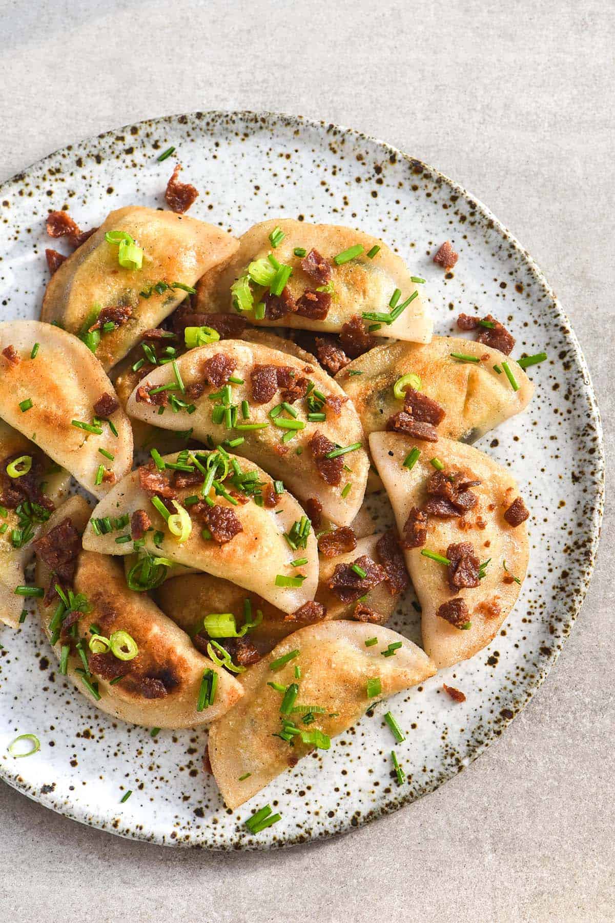 An aerial image of a white speckled ceramic plate topped with gluten free pierogi. The pierogi are sprinkled with chopped vegetarian bacon and chives. The plate sits on a beige stone backdrop