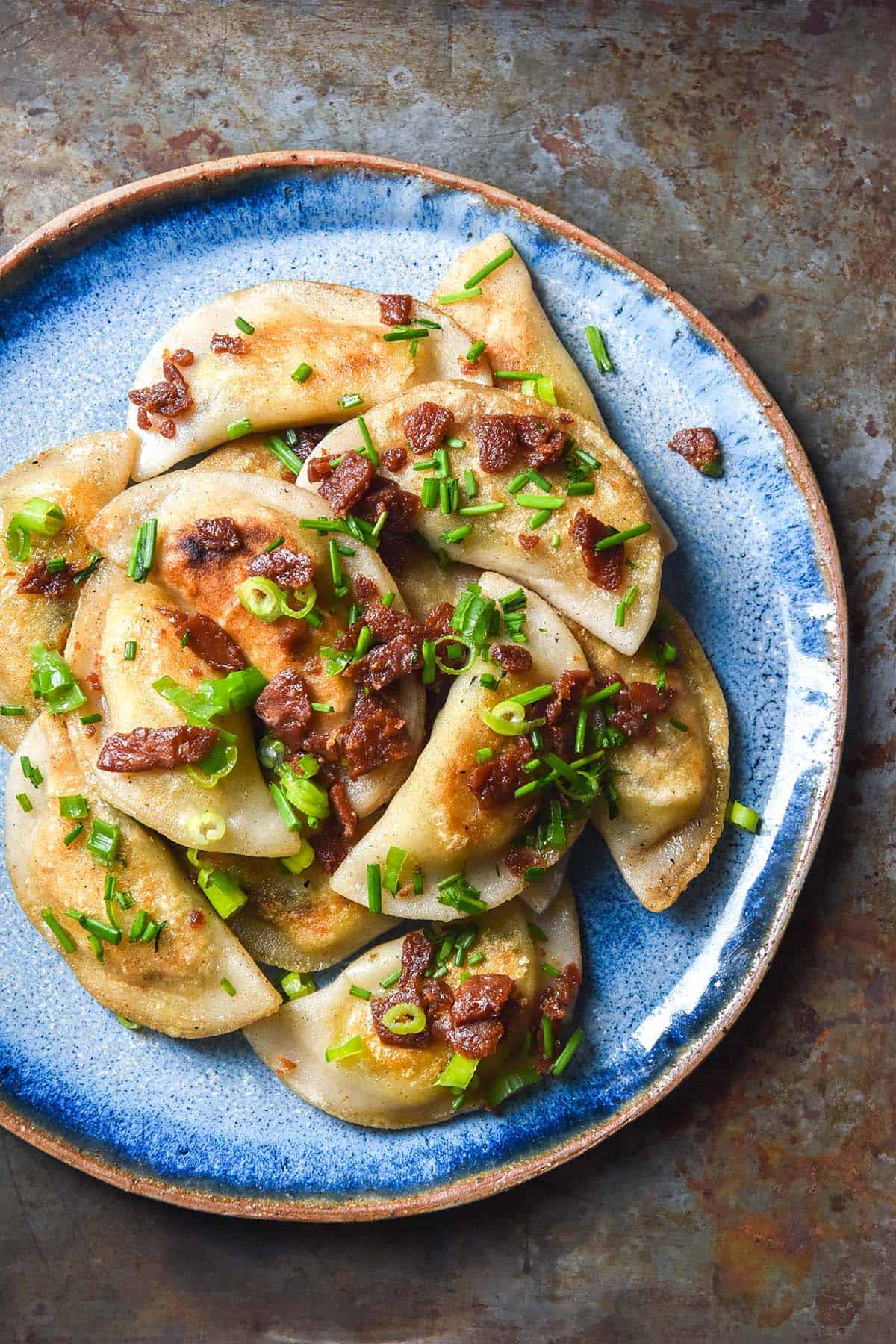 An aerial image of gluten free pierogi on a bright blue ceramic plate. The pierogi are topped with vegetarian bacon, chopped chives and spring onion greens. The plate sits atop a medium blue steel backdrop.