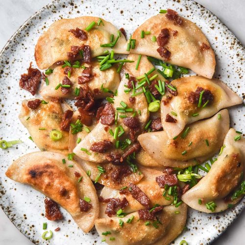 An aerial close up image of a white ceramic plate topped with gluten free pierogi. The pierogi are finished with vegetarian bacon and chives and the plate sits atop a white marble table.