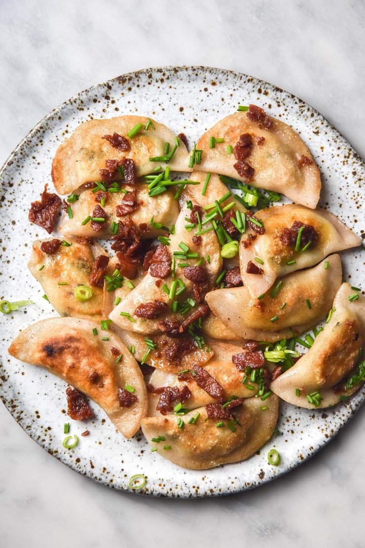 An aerial close up image of a white ceramic plate topped with gluten free pierogi. The pierogi are finished with vegetarian bacon and chives and the plate sits atop a white marble table.