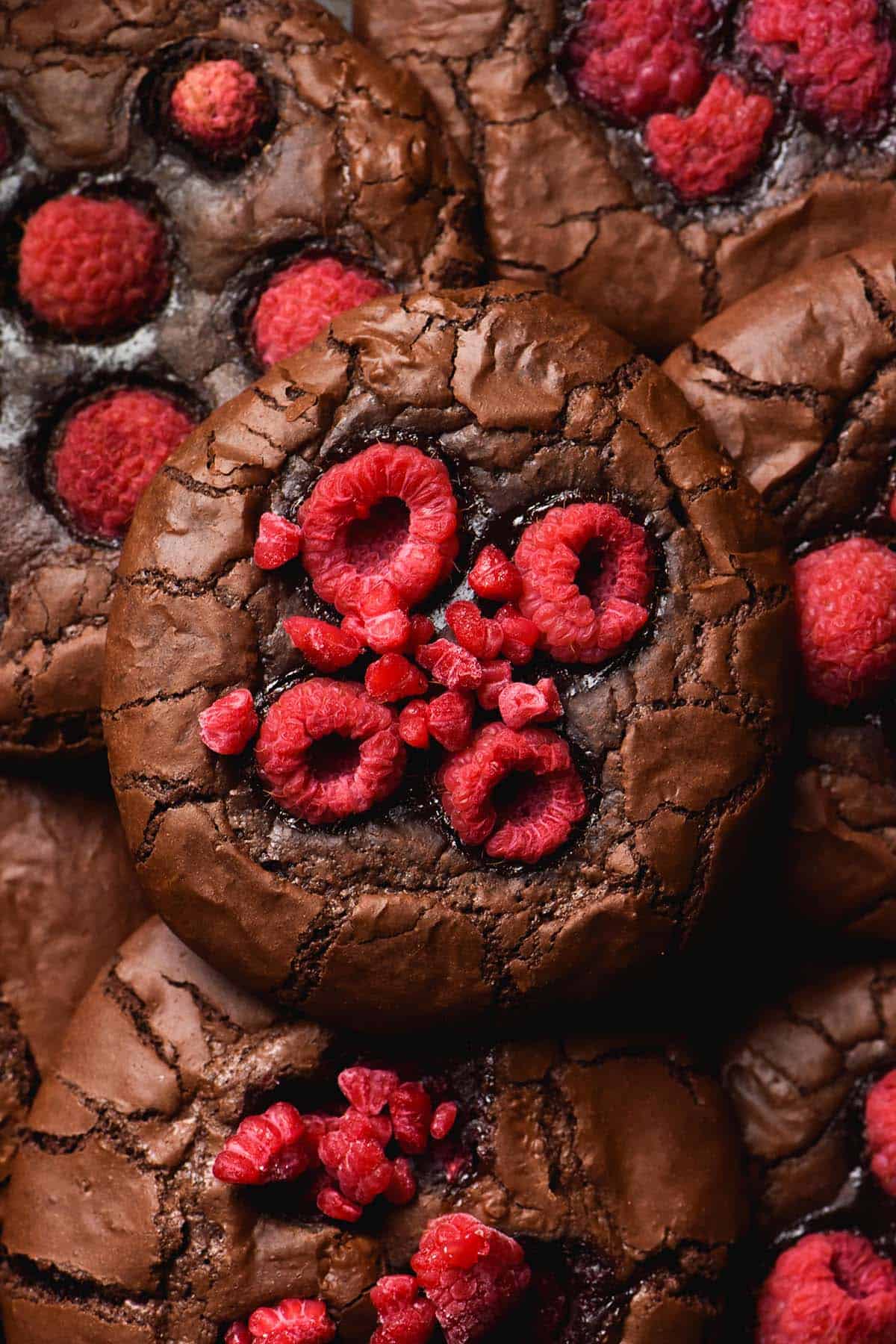 An aerial image of a pile of raspberry brownie cookies. The central cookie is topped with lots of raspberries and the cookie is molten around the brownie edges. 