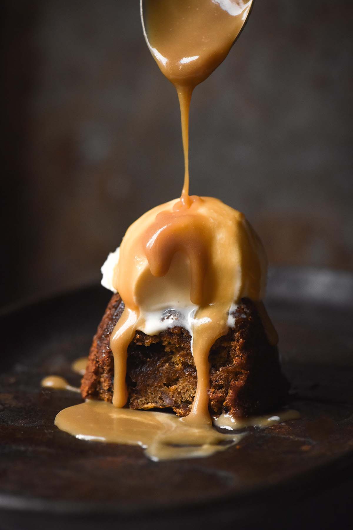 A side on moody image of a gluten free sticky date pudding on a dark grey steel sizzle platter against a dark backdrop. The pudding is topped with a big scoop of vanilla ice cream and butterscotch sauce drizzles onto the pudding from a spoon above.