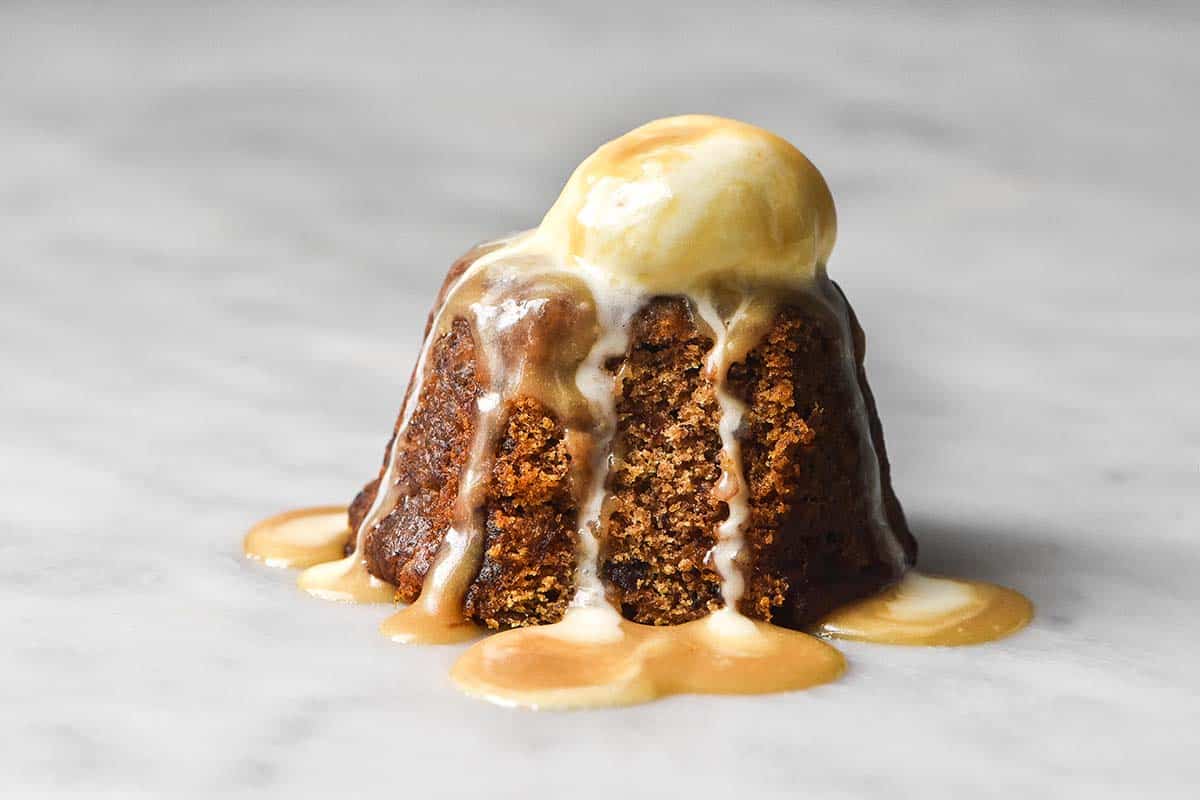 A light and bright image of a gluten free sticky date pudding on a white marble table topped with melting vanilla ice cream and butterscotch sauce