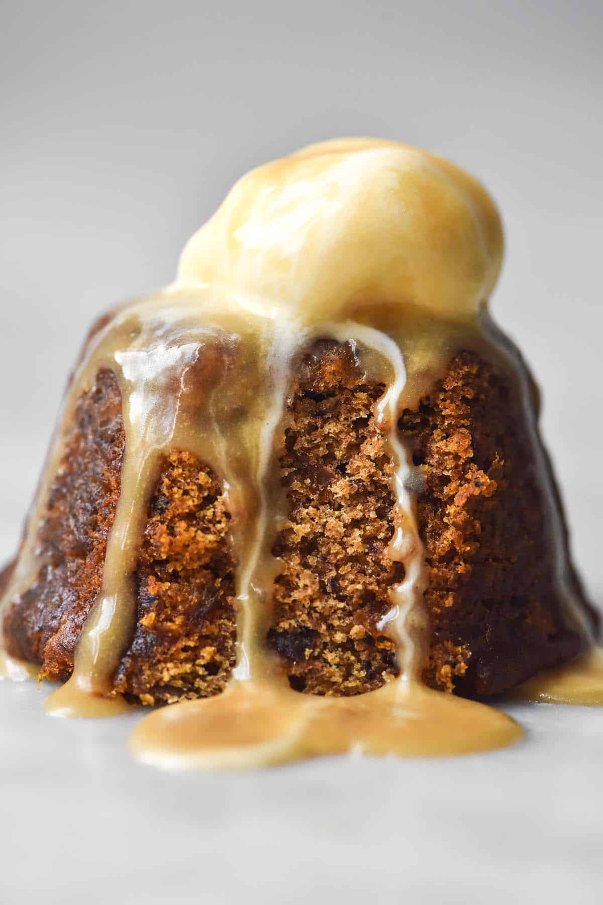 A side on image of a gluten free sticky date pudding on a white marble table. The pudding has been topped with vanilla ice cream and drizzled with butterscotch sauce.