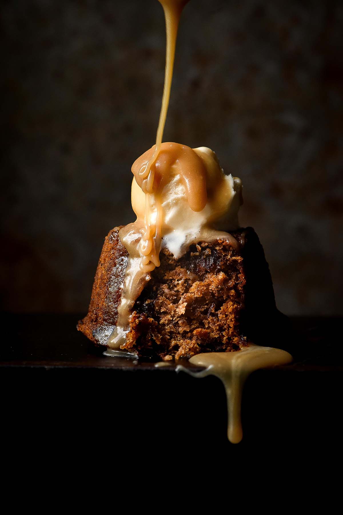 A side on moody image of a gluten free sticky date pudding topped with melting vanilla ice cream. A spoon drizzles butterscotch sauce down onto the pudding from above.