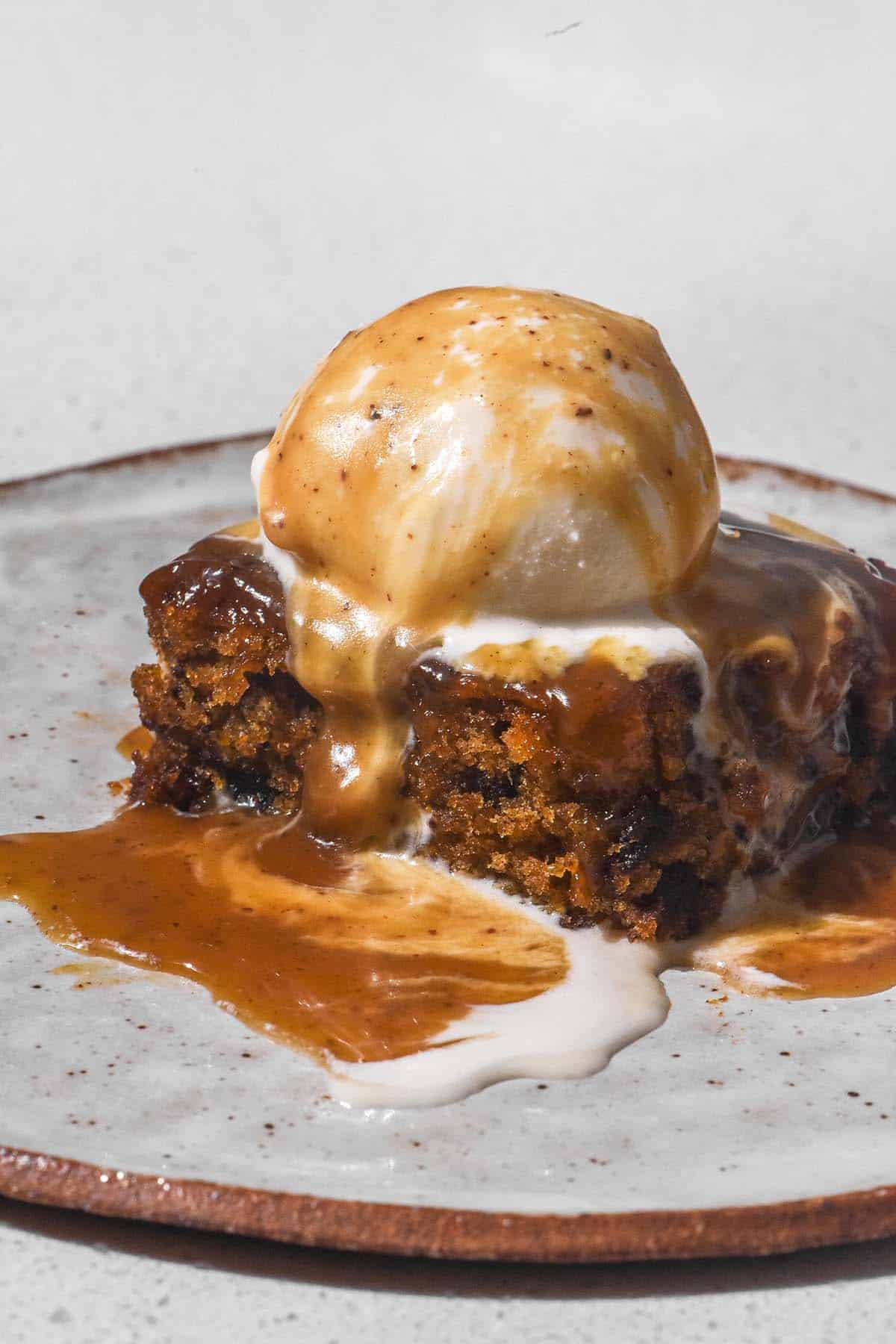 A side on image of a slice of sticky date pudding topped with vanilla ice cream and brown butter butterscotch sauce. The pudding sits on a white ceramic plate atop a white bench top. 
