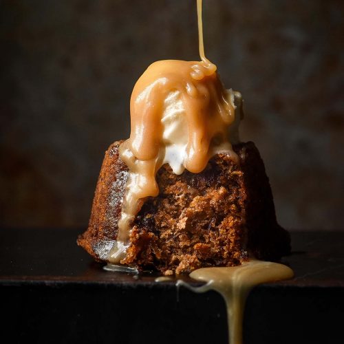 A side on moody image of a gluten free sticky date pudding topped with melting vanilla ice cream. A spoon drizzles butterscotch sauce down onto the pudding from above.