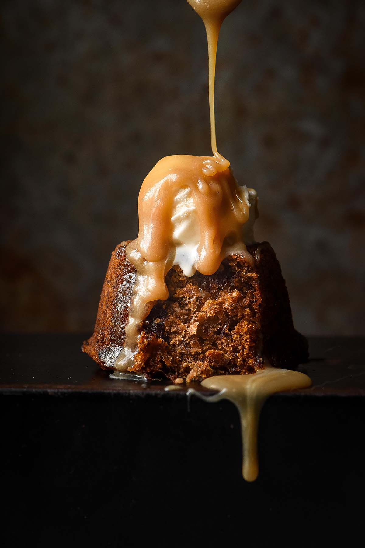 A side on moody image of a gluten free sticky date pudding topped with melting vanilla ice cream. A spoon drizzles butterscotch sauce down onto the pudding from above.