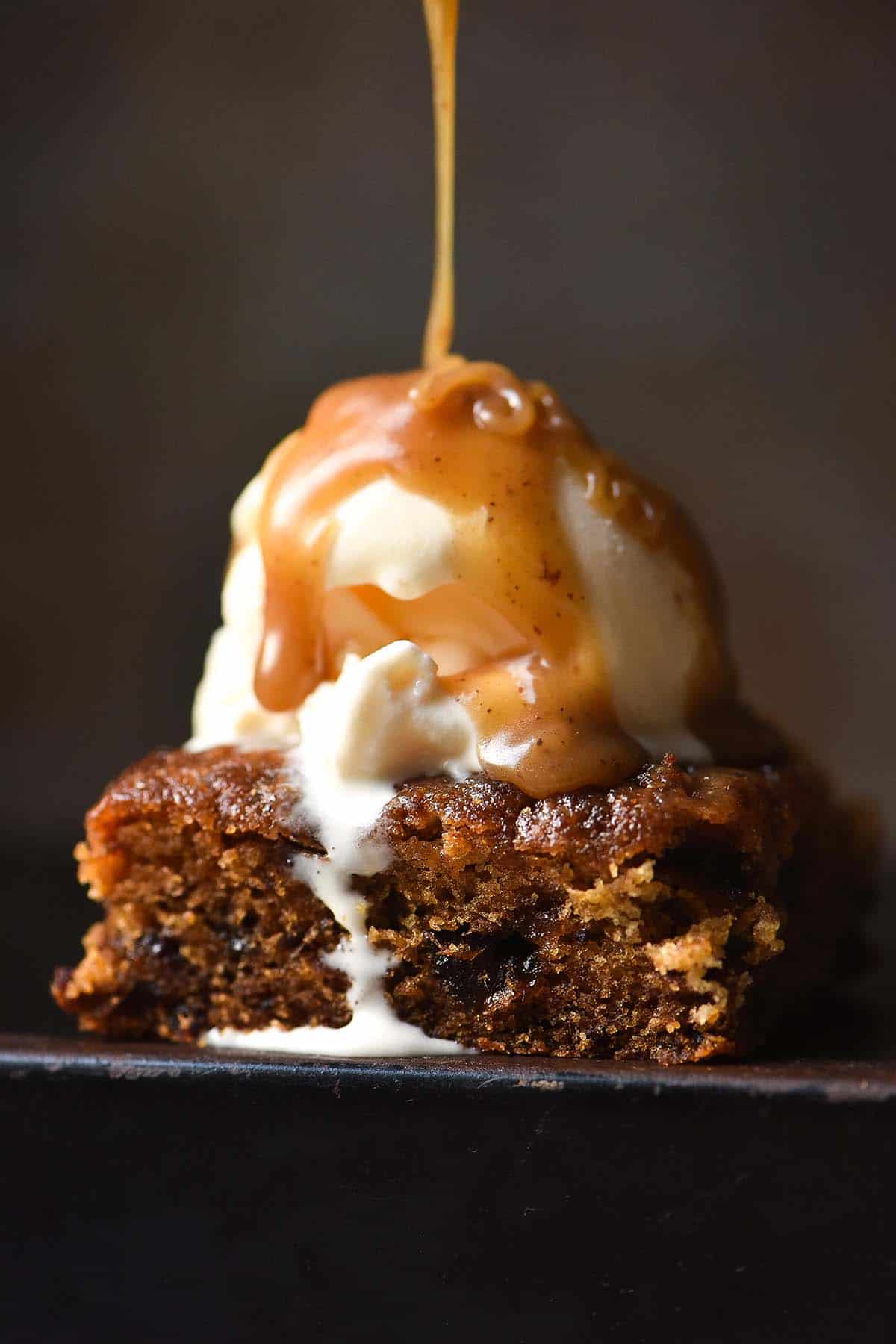 A dark and moody image of a slice of sticky date pudding against a dark backdrop topped with vanilla ice cream and finished with a drizzle of butterscotch sauce