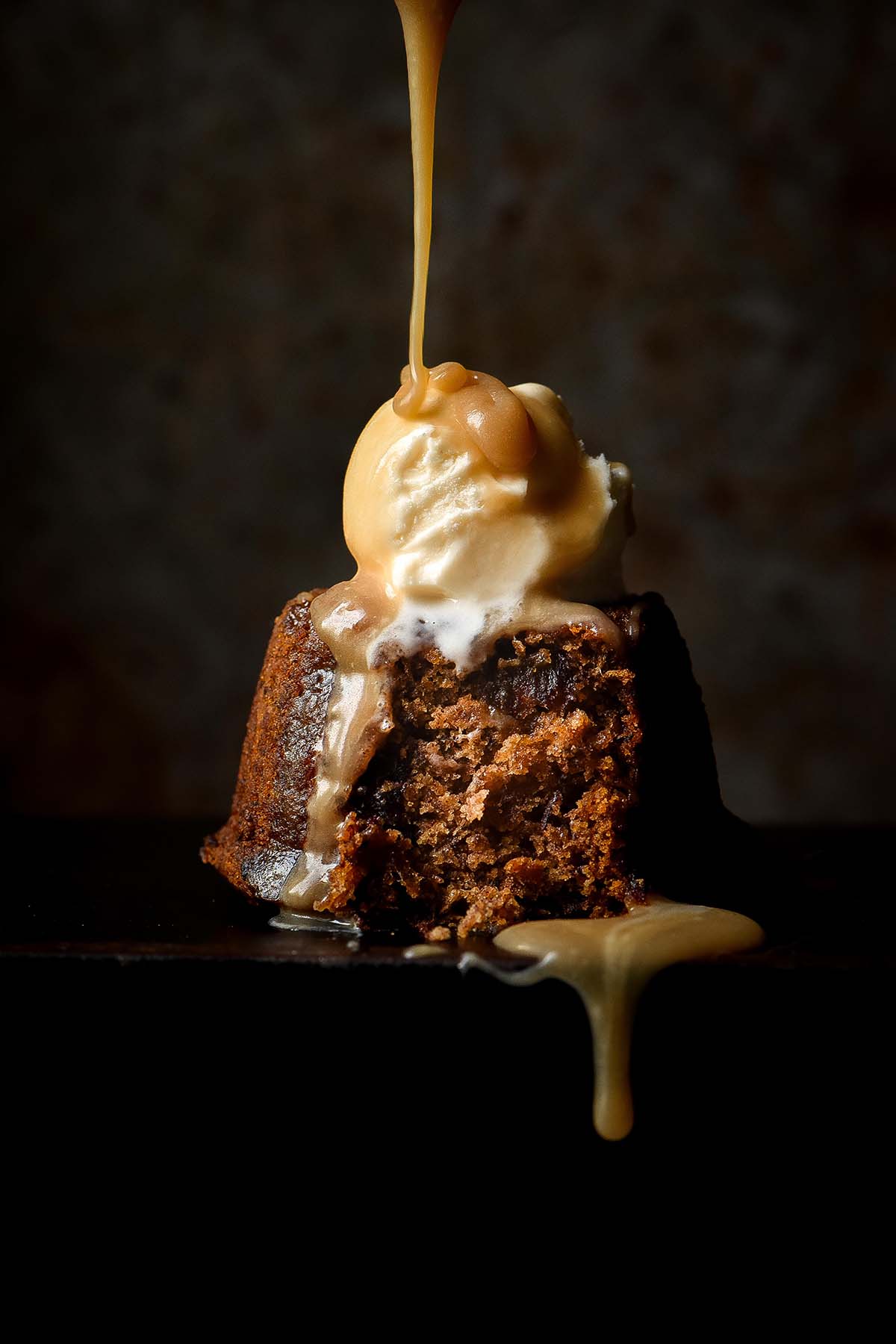 A dark and moody side on image of a gluten free sticky date pudding on a black plate against a dark steel backdrop. The pudding is topped with vanilla ice cream which is being drizzled with butterscotch sauce from above.