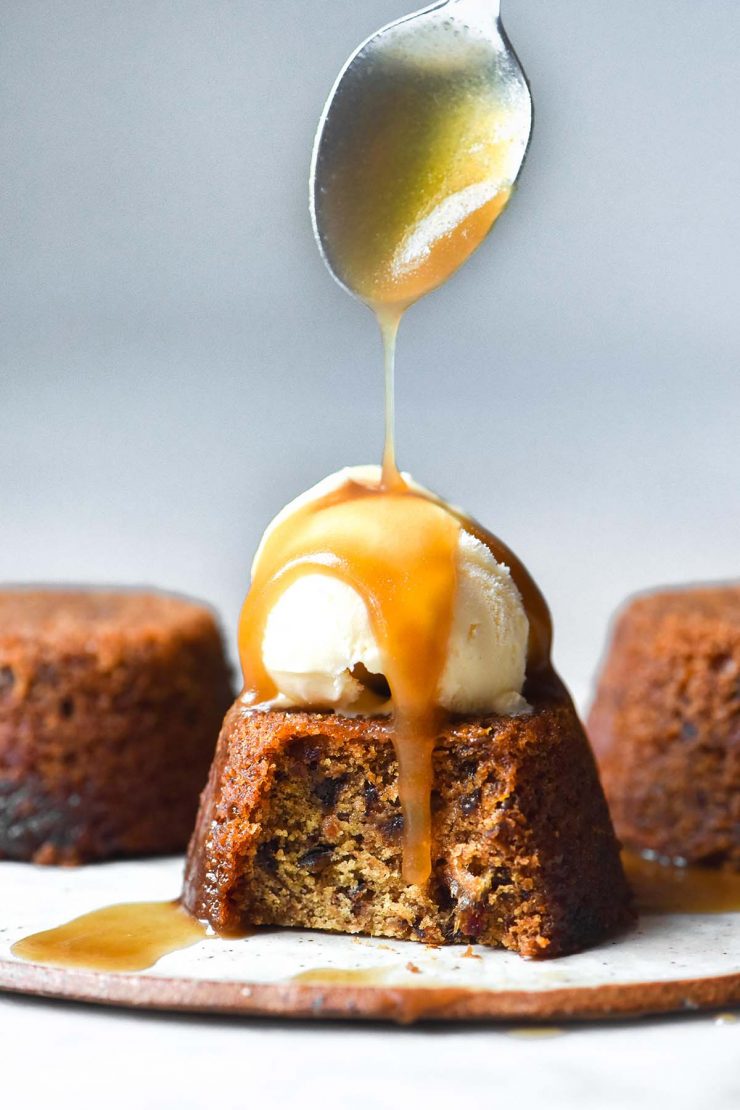 A side on image of a gluten free vegan sticky toffee pudding on a white ceramic plate against a white backdrop. The pudding is topped with vanilla ice cream and a spoon extends from the top of the image to drizzle butterscotch sauce over the top