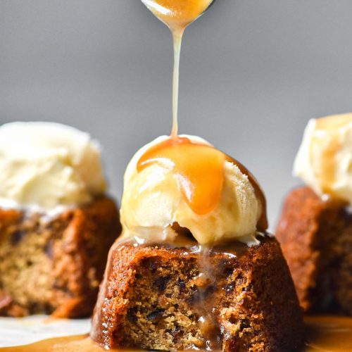 A side on image of three gluten free vegan sticky date puddings on a white plate against a white backdrop. The puddings have all had a spoonful taken, revealing the inside of the pudding. They are topped with vanilla ice cream and the central pudding is being drizzled with vegan butterscotch sauce.