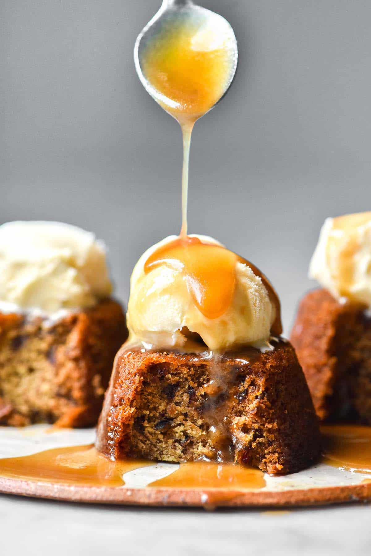 A side on image of three gluten free vegan sticky date puddings on a white plate against a white backdrop. The puddings have all had a spoonful taken, revealing the inside of the pudding. They are topped with vanilla ice cream and the central pudding is being drizzled with vegan butterscotch sauce. 