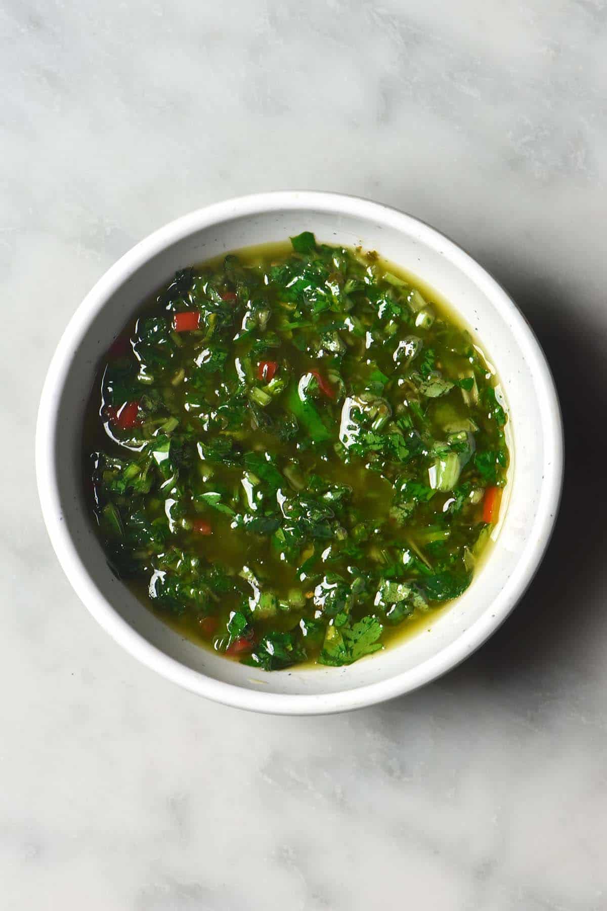 An aerial image of a small white bowl filled with low FODMAP chimichurri. The bowl sits atop a white marble table. 