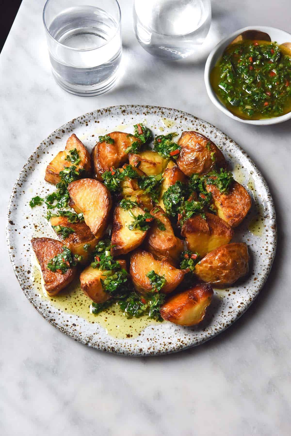 An aerial image of a white speckled ceramic plate topped with crispy potatoes smothered in low FODMAP chimichurri. The plate sits on a white marble table and water glasses plus a small bowl of chimichurri sit to the top of the plate. 