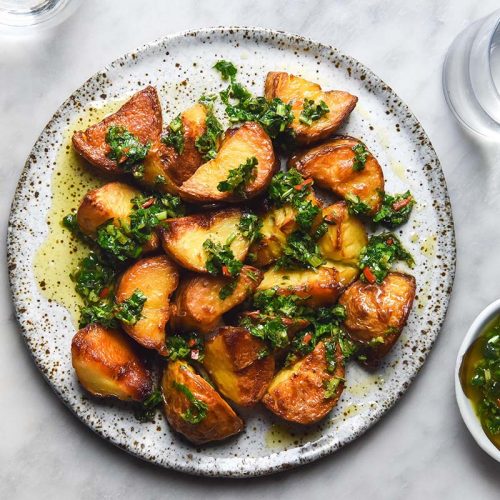 An aerial image of a white speckled ceramic plate topped with crispy potatoes smothered in low FODMAP chimichurri. The plate sits on a white marble table and water glasses plus a small bowl of chimichurri sit to the right hand side of the plate.