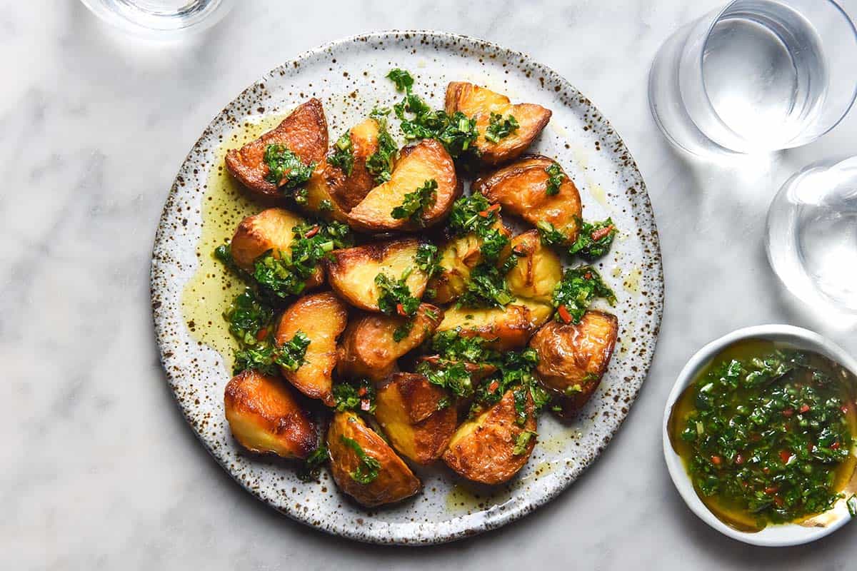 An aerial image of a white speckled ceramic plate topped with crispy potatoes smothered in low FODMAP chimichurri. The plate sits on a white marble table and water glasses plus a small bowl of chimichurri sit to the right hand side of the plate. 