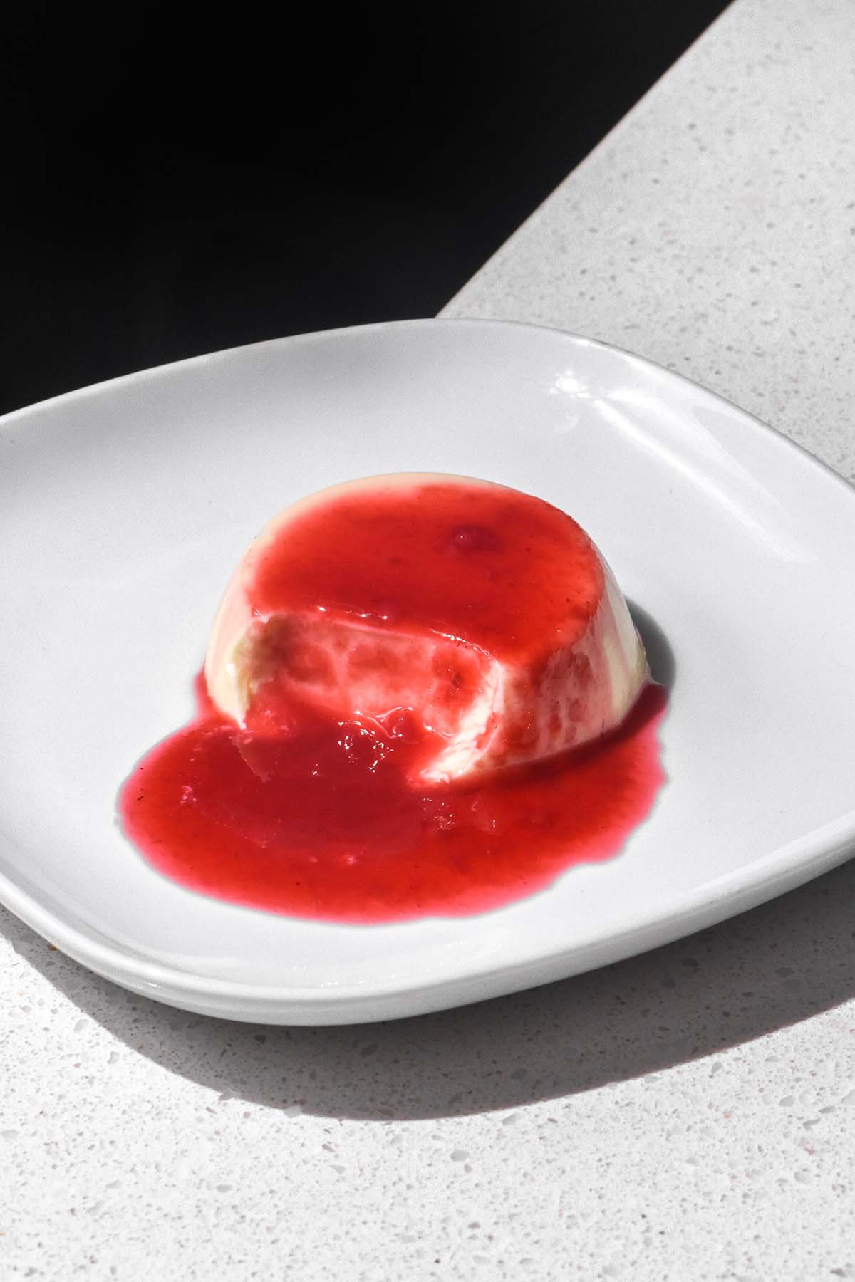 A side on image of a panna cotta topped with raspberry coulis on a white plate against a contrasting black and white backdrop. A spoon has been taken from the panna cotta, revealing the creamy insides. 