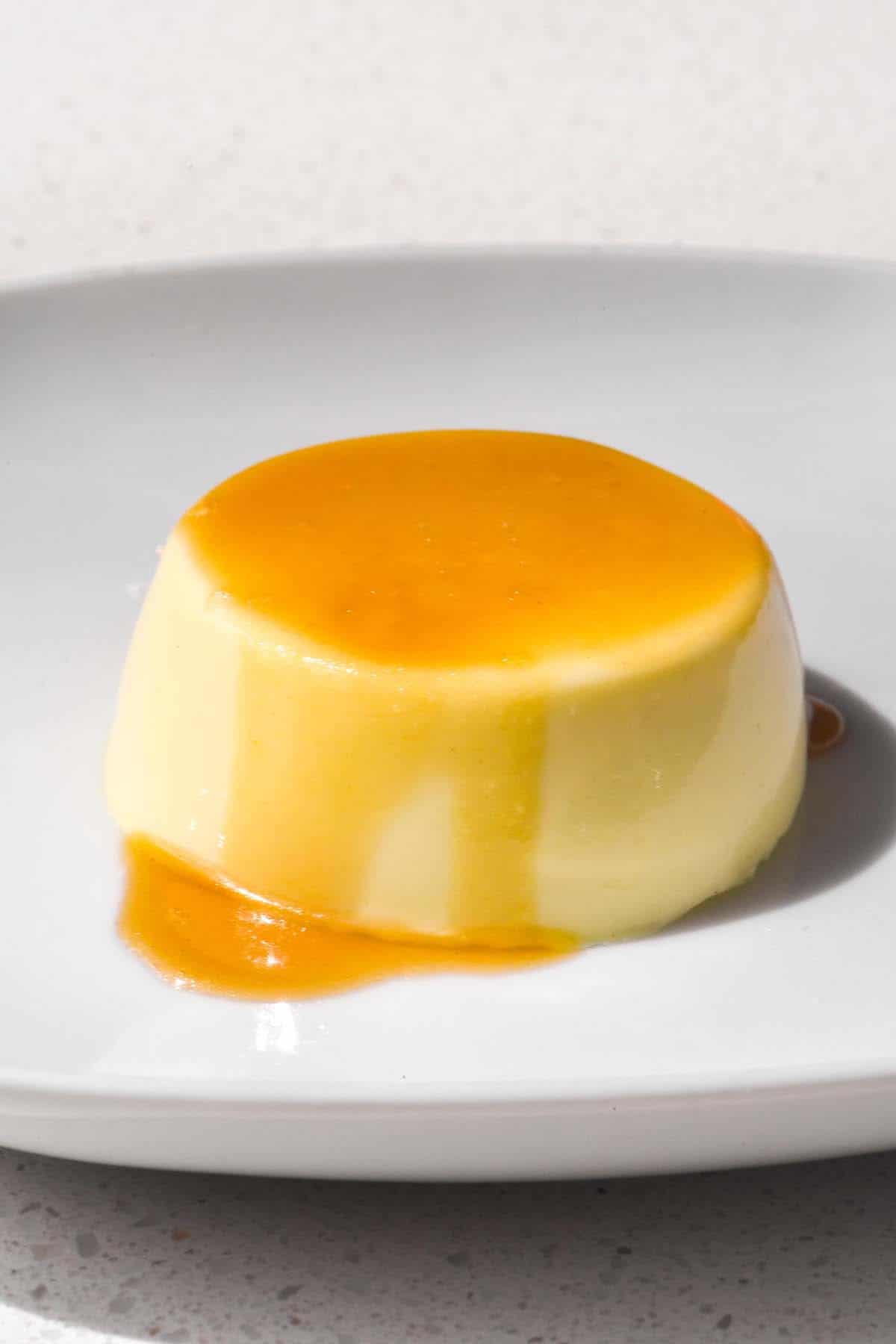 A side on image of a panna cotta with agar agar on a white plate against a white stone benchtop. The panna cotta has been drizzled with butterscotch sauce and sprinkled with sea salt flakes. 