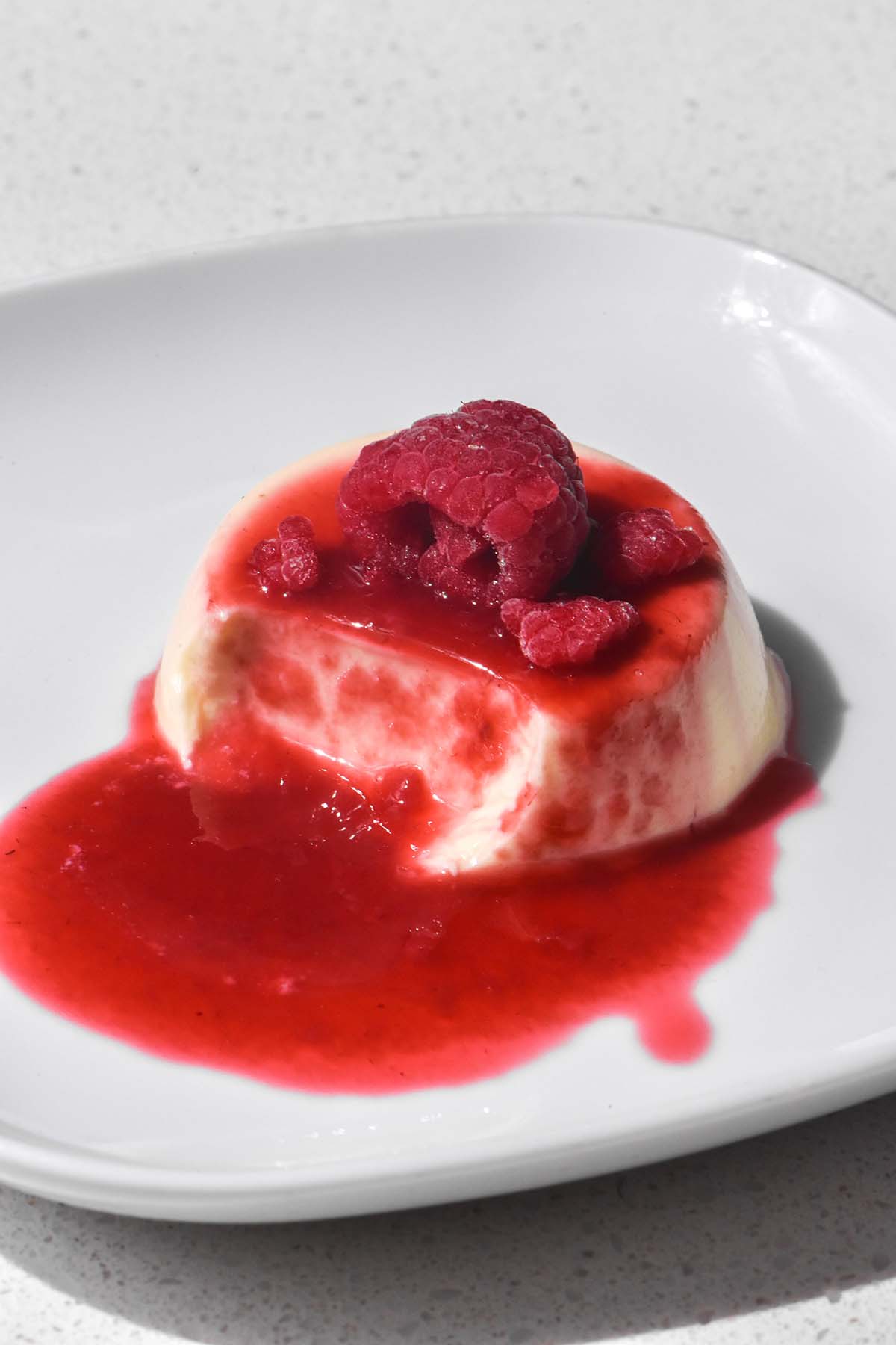 A close up image of a panna cotta on a white plate topped with raspberry coulis and whole raspberries. A spoon has been taken from the panna cotta revealing the creamy insides. 