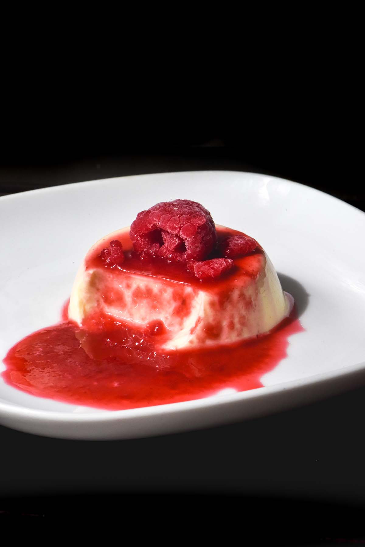 A dark and moody shot of a panna cotta on a small white plate topped with raspberry coulis and extra raspberries. 