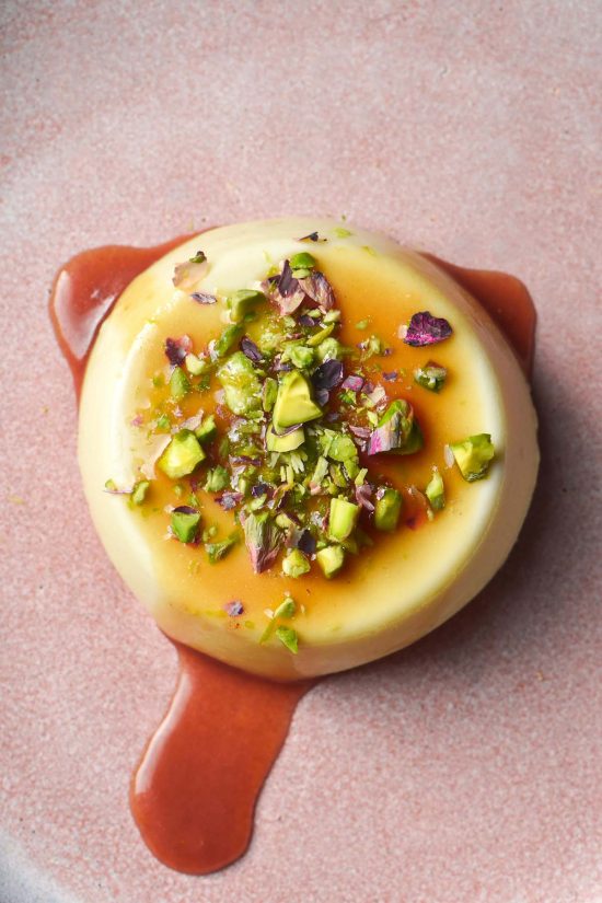 An aerial image of a panna cotta on a pale pink ceramic plate topped with chopped pistachios and a vibrant caramel sauce