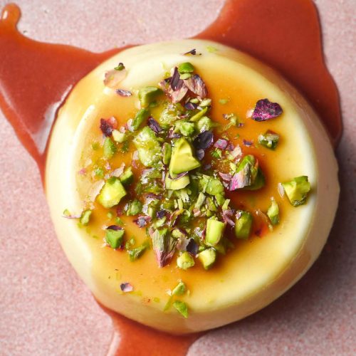 An aerial image of a panna cotta on a pale pink ceramic plate topped with chopped pistachios and a vibrant caramel sauce