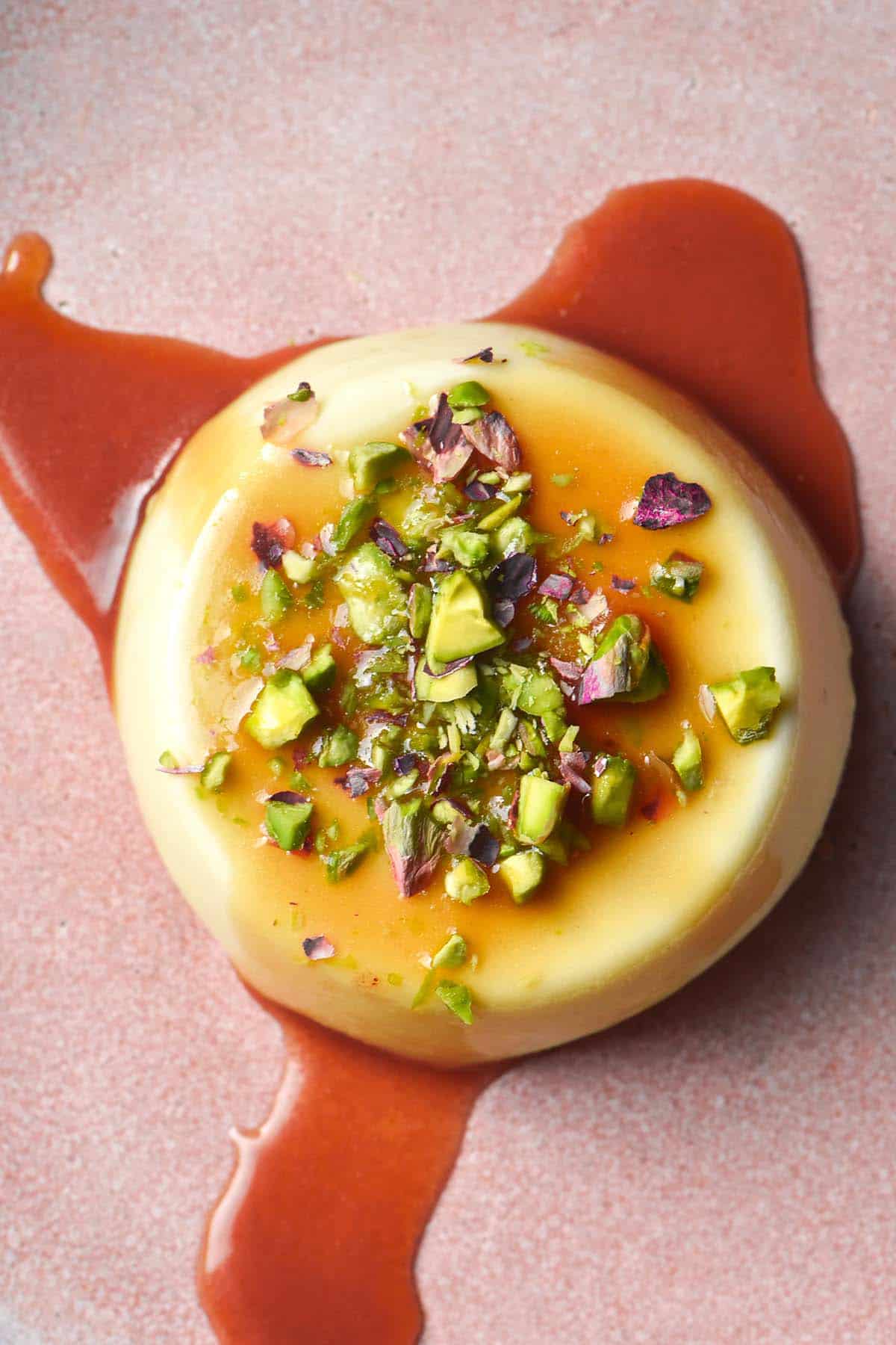 An aerial image of a panna cotta on a pale pink ceramic plate topped with chopped pistachios and a vibrant caramel sauce. 