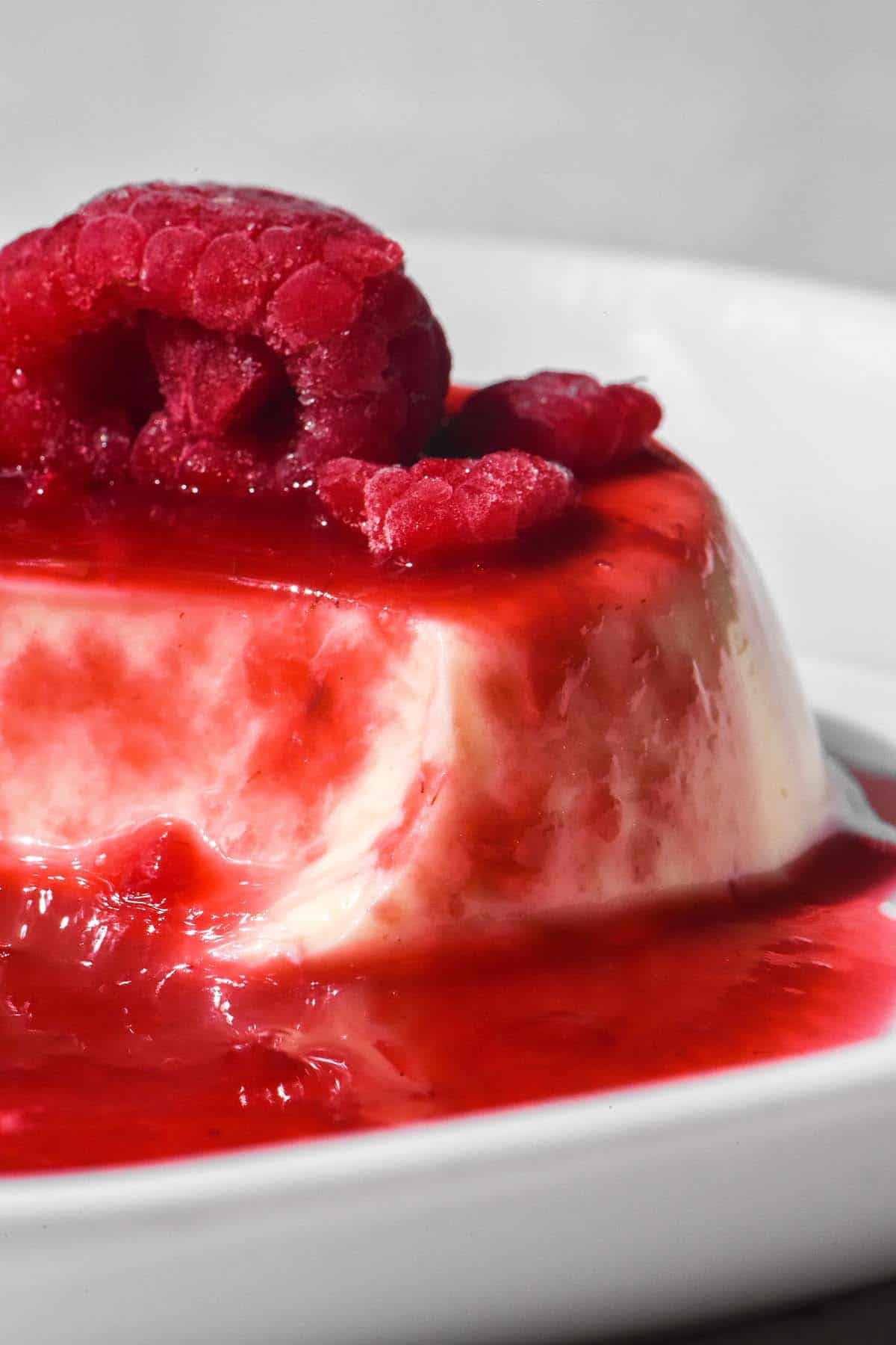 A close up side on image of a panna cotta topped with raspberry coulis and a frozen raspberry. The panna cotta sits atop a white plate against a white backdrop. 