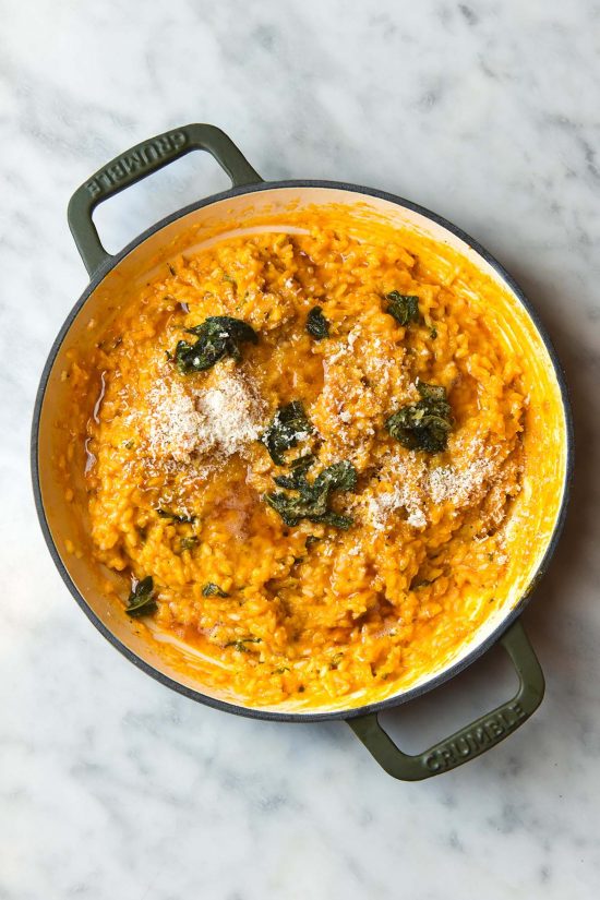 An aerial view of a skillet of pumpkin risotto on a white marble table. The pumpkin risotto is topped with brown sage butter and finely grated parmesan