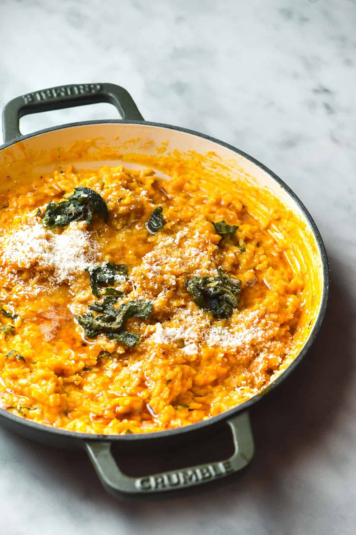 An image of a dark olive skillet filled with pumpkin risotto on a white marble table. The risotto is topped with brown sage butter and parmesan. 
