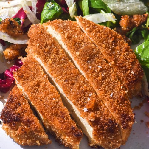 A brightly lit close up image of gluten free vegan schnitzel sliced on a white plate surrounded by salad.