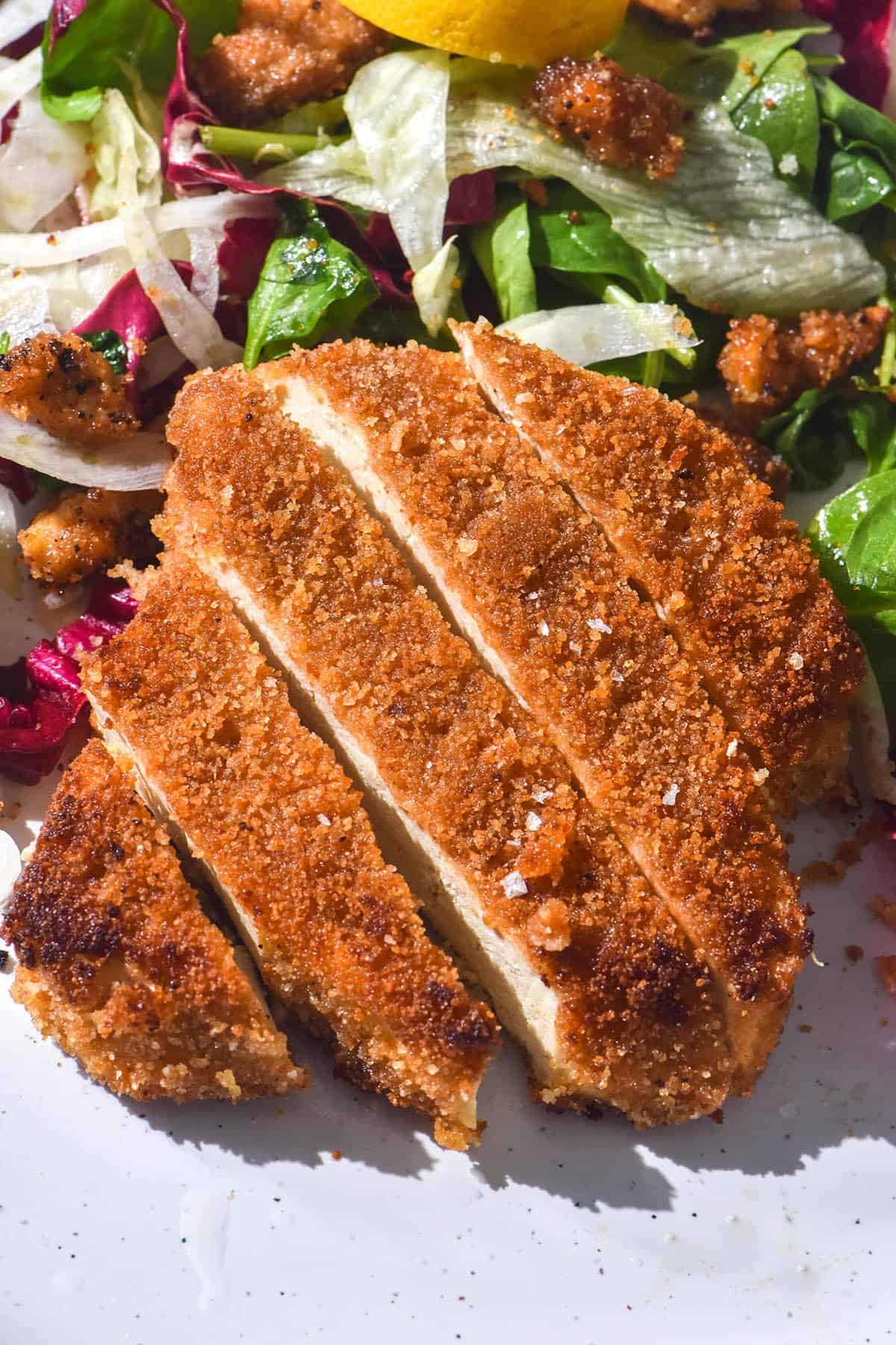 A brightly lit side on image of a gluten free vegan schnitzel sliced into fingers on a white plate. A dressed salad and lemon wedge sit in the background of the image. 