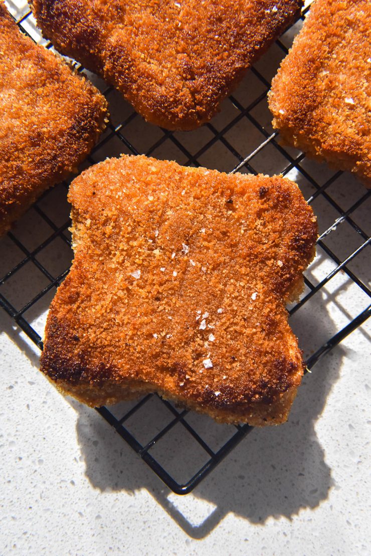 A brightly lit aerial image of gluten free vegan schnitzels on a cooling rack atop a white stone bench top. The schnitzels are golden brown and topped with a small sprinkle of sea salt flakes.