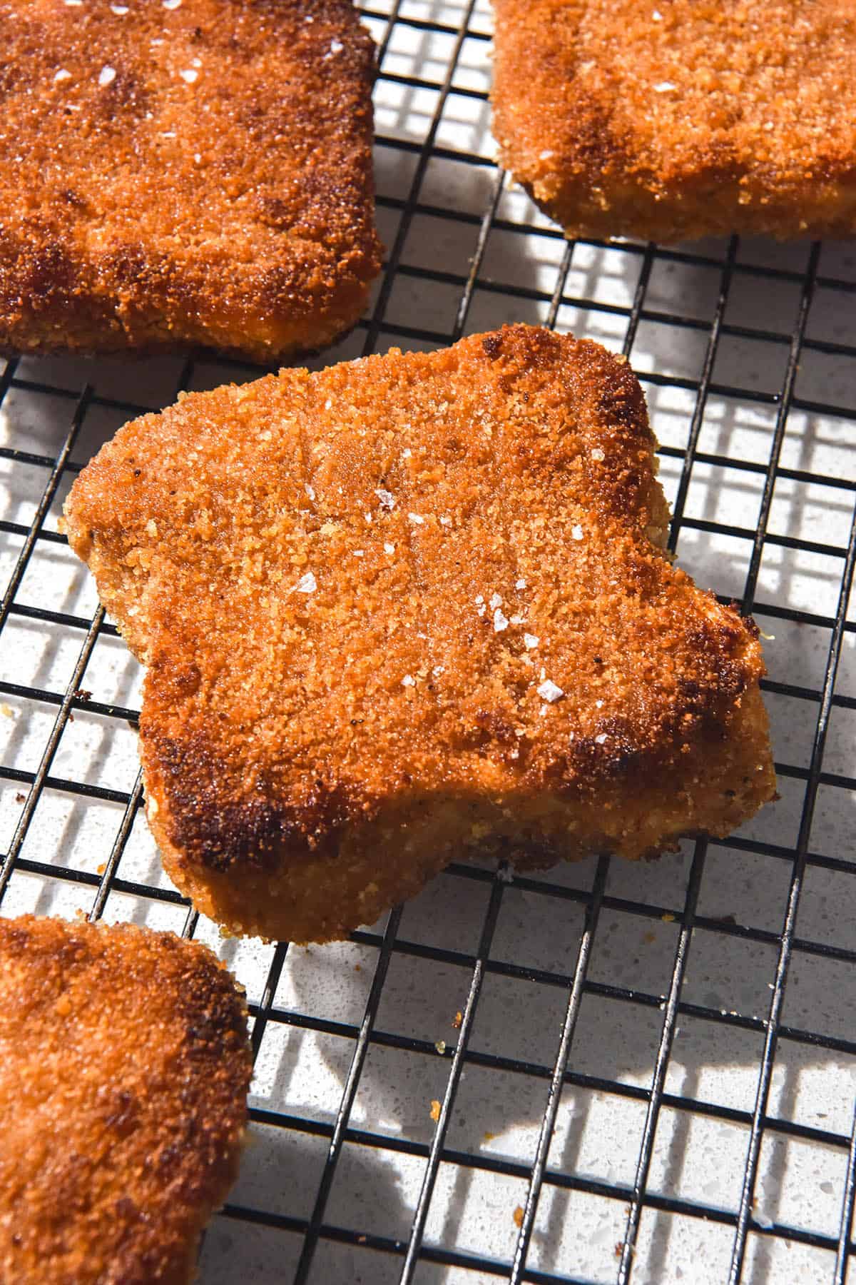 A brightly lit side on image of gluten free vegan schnitzels on a cooling rack atop a white stone bench top. The schnitzels are golden brown and topped with a small sprinkle of sea salt flakes. 