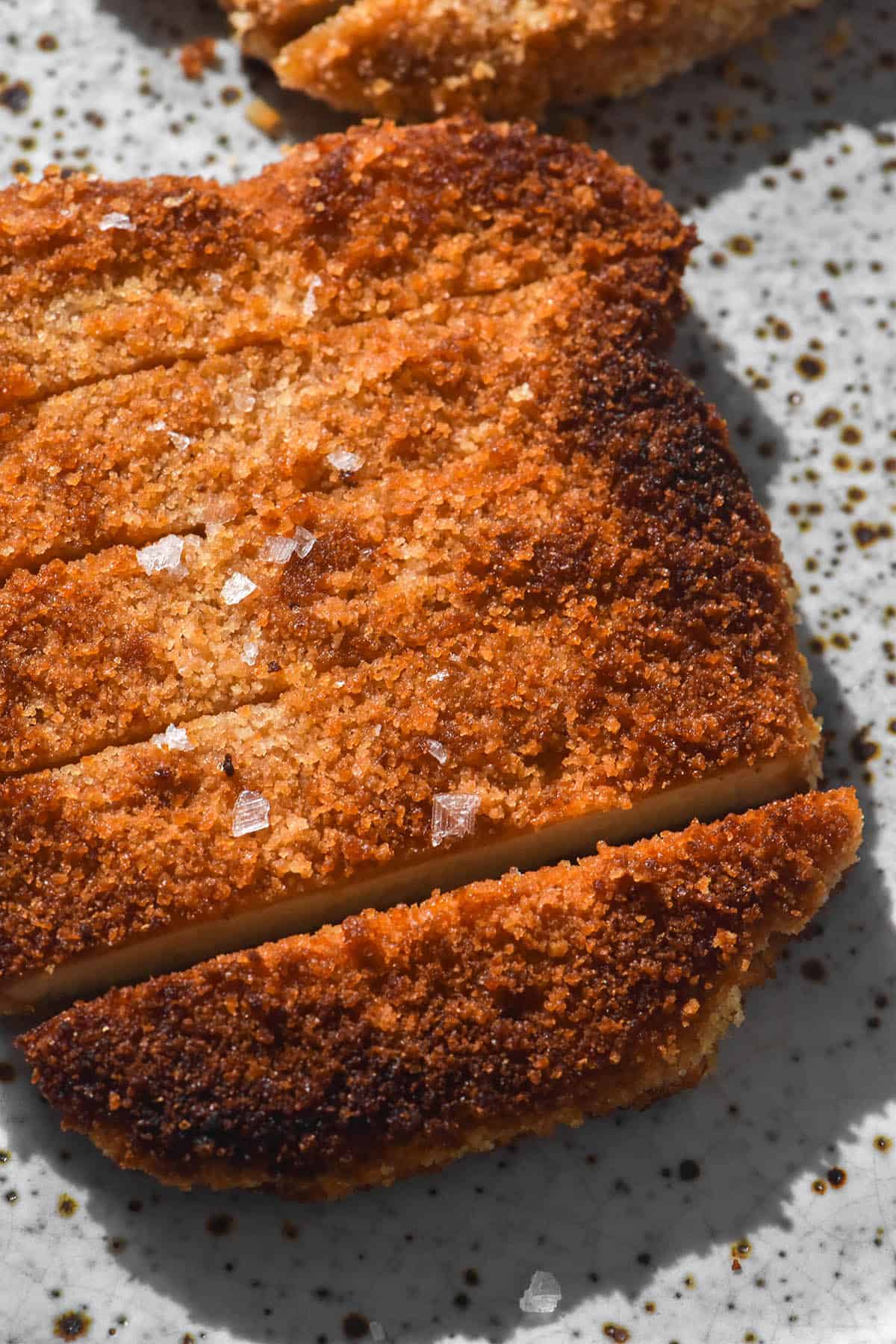 A close up aerial image of a gluten free vegan schnitzel that has been sliced and topped with some sea salt flakes. The schnitzel sits atop a white speckled ceramic plate in bright lighting. 