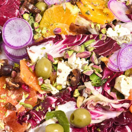 A close up macro image of a winter citrus salad in bright light. The salad contains oranges, radicchio, fennel, watermelon radishes, olives and nuts which are mixed together for a rainbow coloured salad.