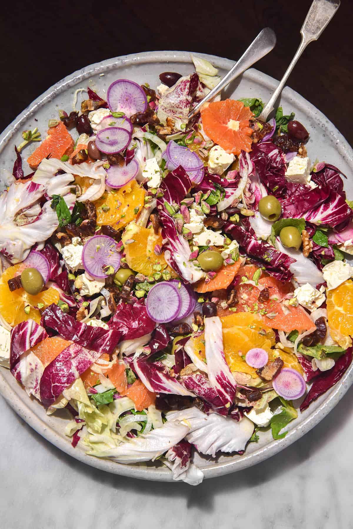 An aerial image of a brightly coloured winter Citrus salad on a large white serving plate. The plate sits half on a white marble table and half off, creating a dark and light effect around the serving platter.