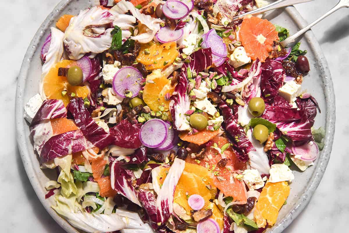 A brightly lit image of a rainbow coloured winter citrus salad on a large white serving platter atop a white marble table. The salad contains oranges, fennel, radicchio, olives, watermelon radishes and nuts which intermingle to form a bright rainbow salad. 