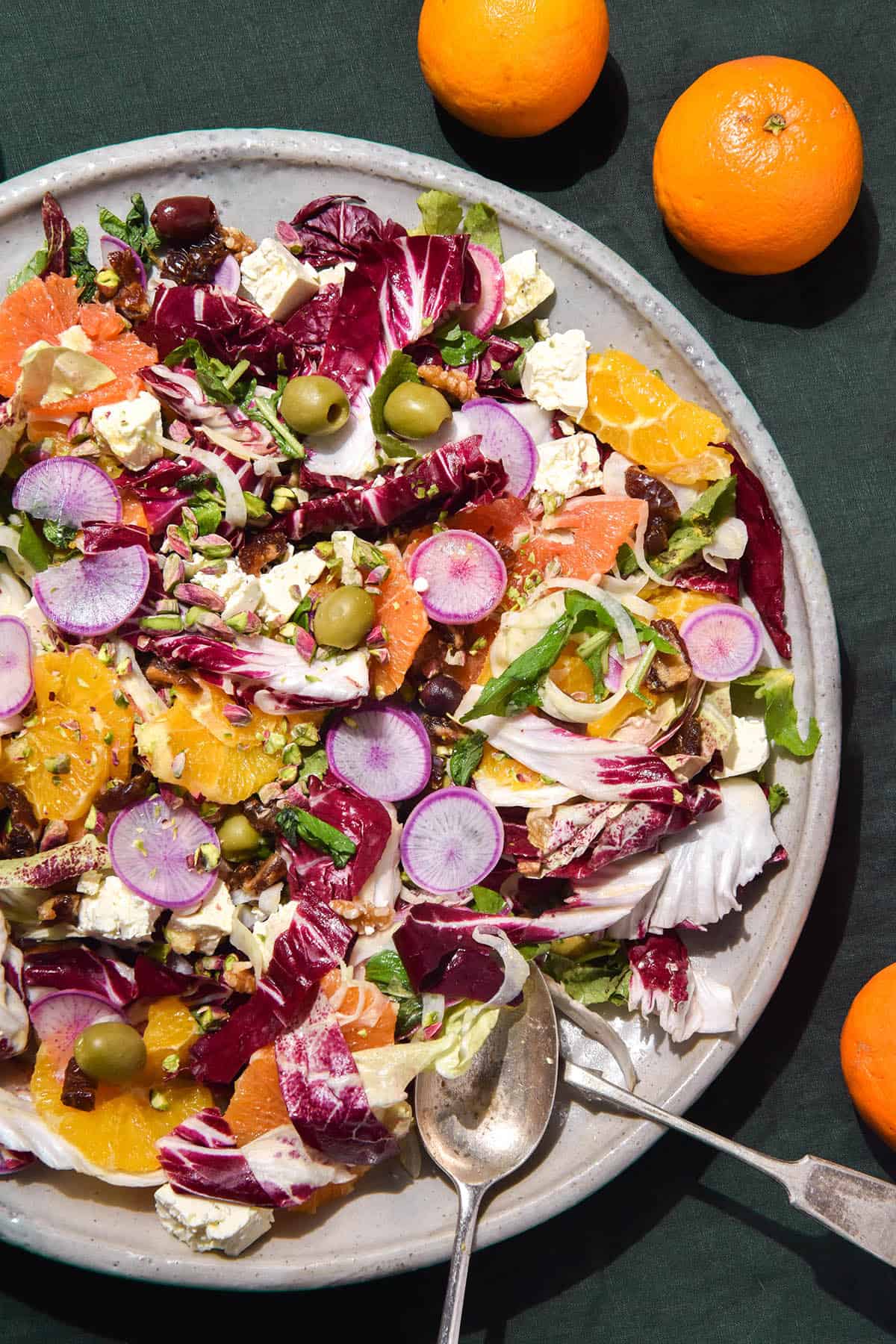 A close up aerial image of a winter citrus salad on a large white serving platter atop an olive green tablecloth. Two silver spoons sit in the salad to the bottom right of the image. 