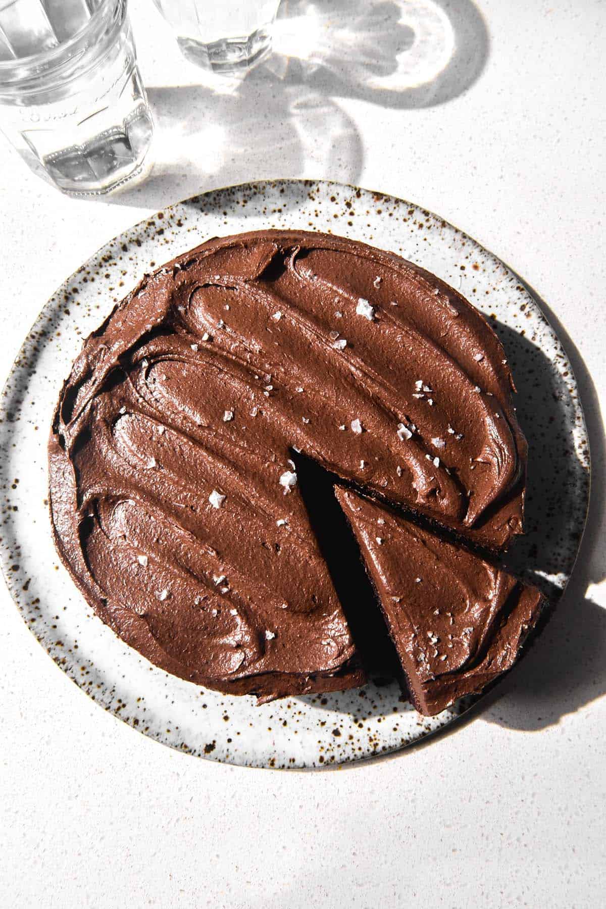 An aerial image of a gluten free vegan buckwheat chocolate cake topped with chocolate buttercream and sea salt flakes. The cakes sits on a white speckled ceramic plate atop a white stone bench top. Two glasses of water create a light and shadow pattern across the top left of the image. 